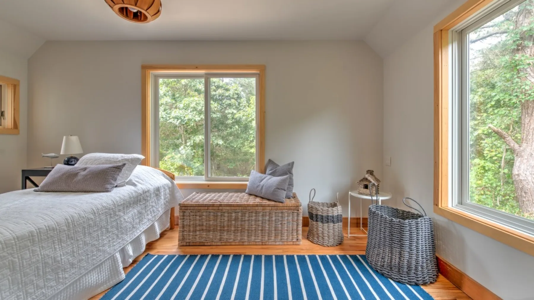Un dormitorio luminoso tiene una cama, una ventana con vista a los árboles y una alfombra azul a rayas.