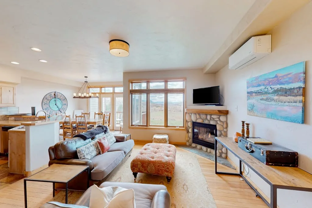 A bright living room opens onto a dining area, with large windows and a fireplace.