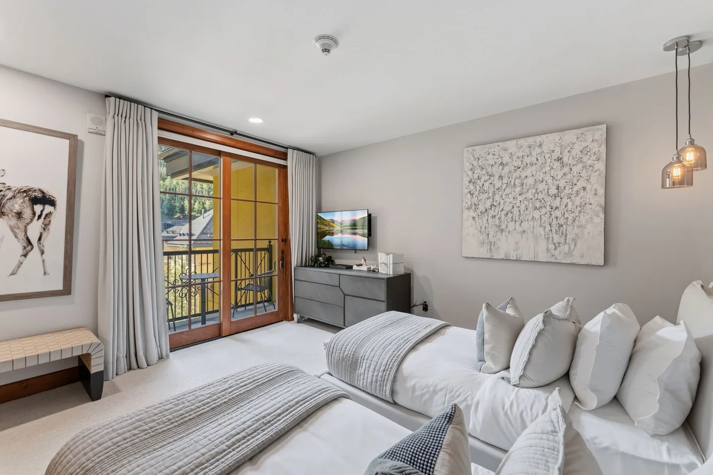 Un dormitorio luminoso con camas, un televisor y puertas de vidrio hacia un balcón.