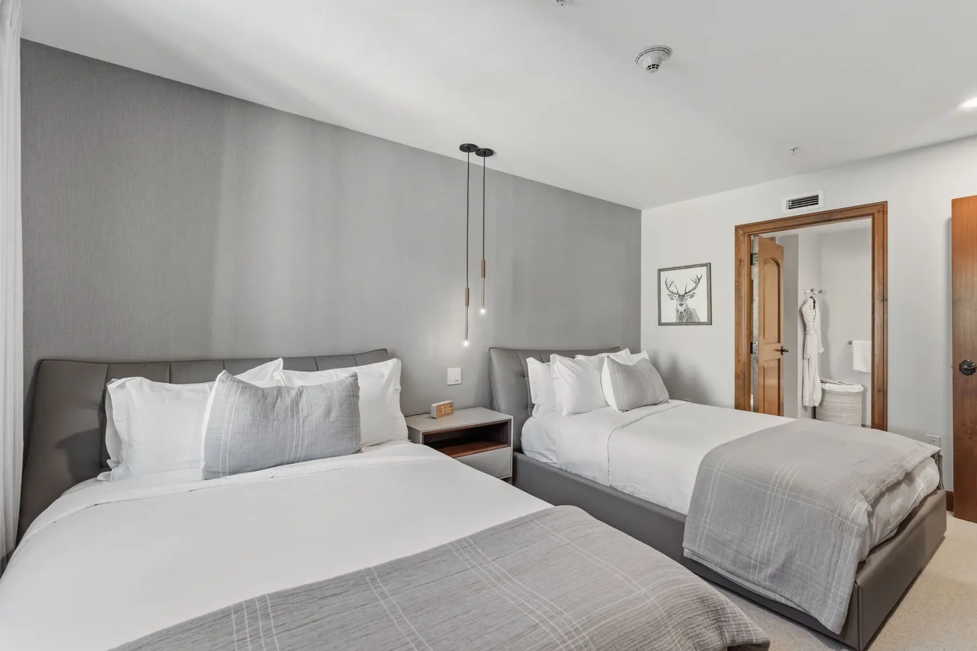 Un dormitorio con dos camas, ropa de cama gris y una puerta al baño.