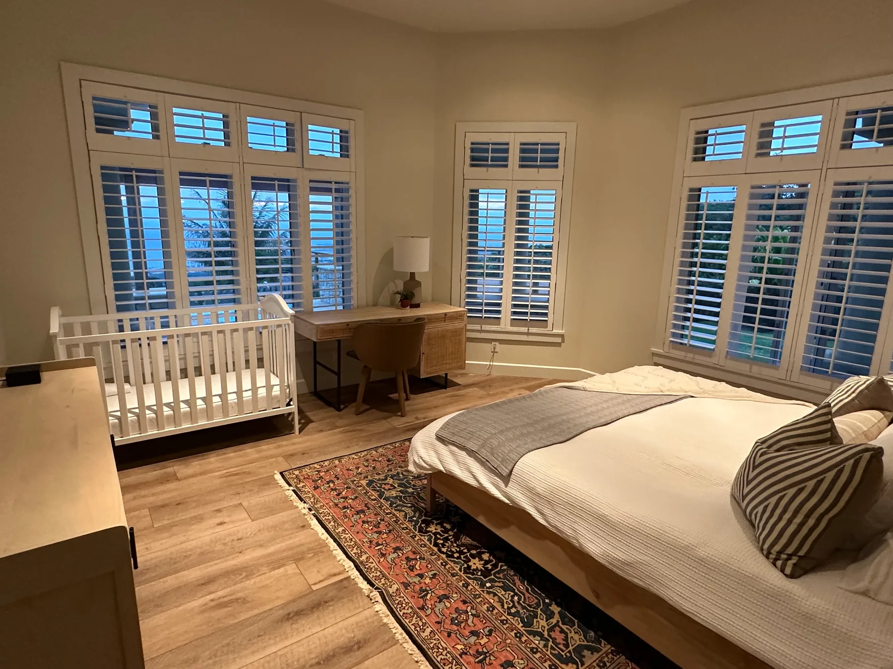 Un dormitorio con una cama, una cuna y un escritorio junto a ventanas con persianas.
