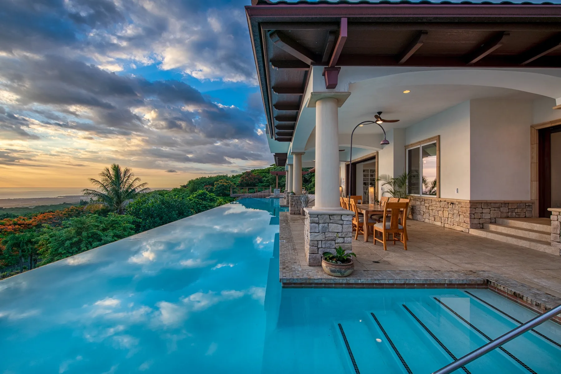 Una piscina al aire libre junto a un patio cubierto mira al océano al atardecer.