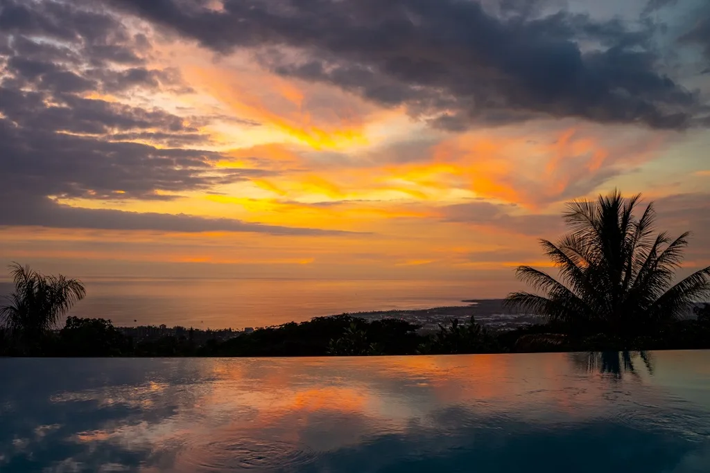 Un atardecer sobre el océano se refleja en una piscina infinita, con palmeras junto a la costa.