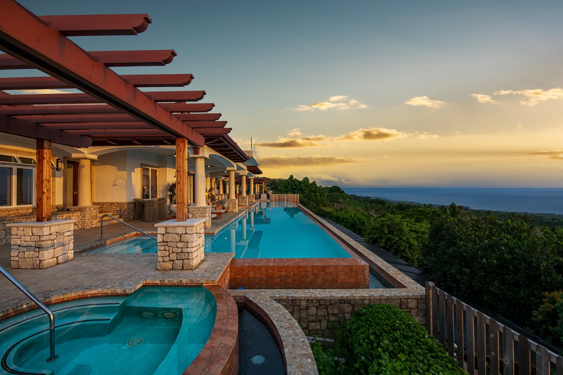 Una piscina junto a una fila de edificios mira hacia la vegetación y el océano al atardecer.