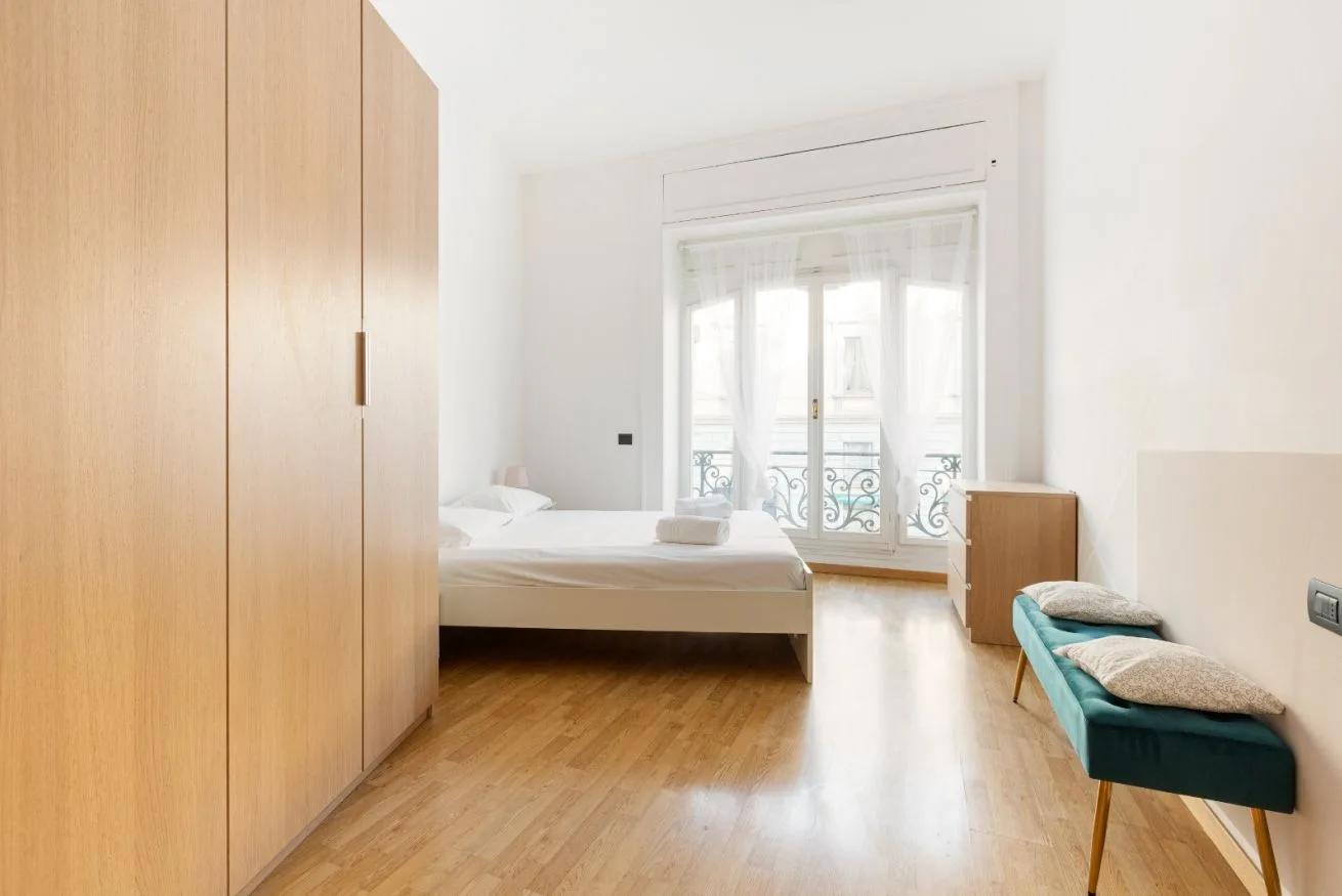 A bright bedroom with a bed beside tall windows and a teal bench.