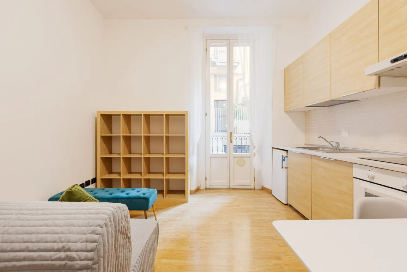 A bright studio room with a bed, open shelving, and a compact kitchen.