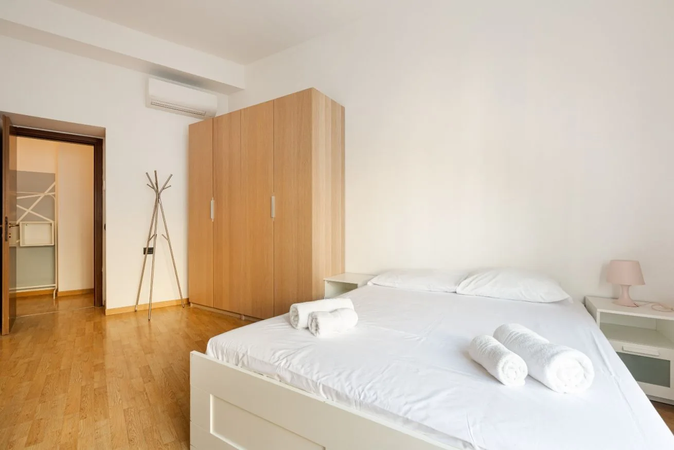 A bright bedroom with a white bed and a wooden wardrobe.