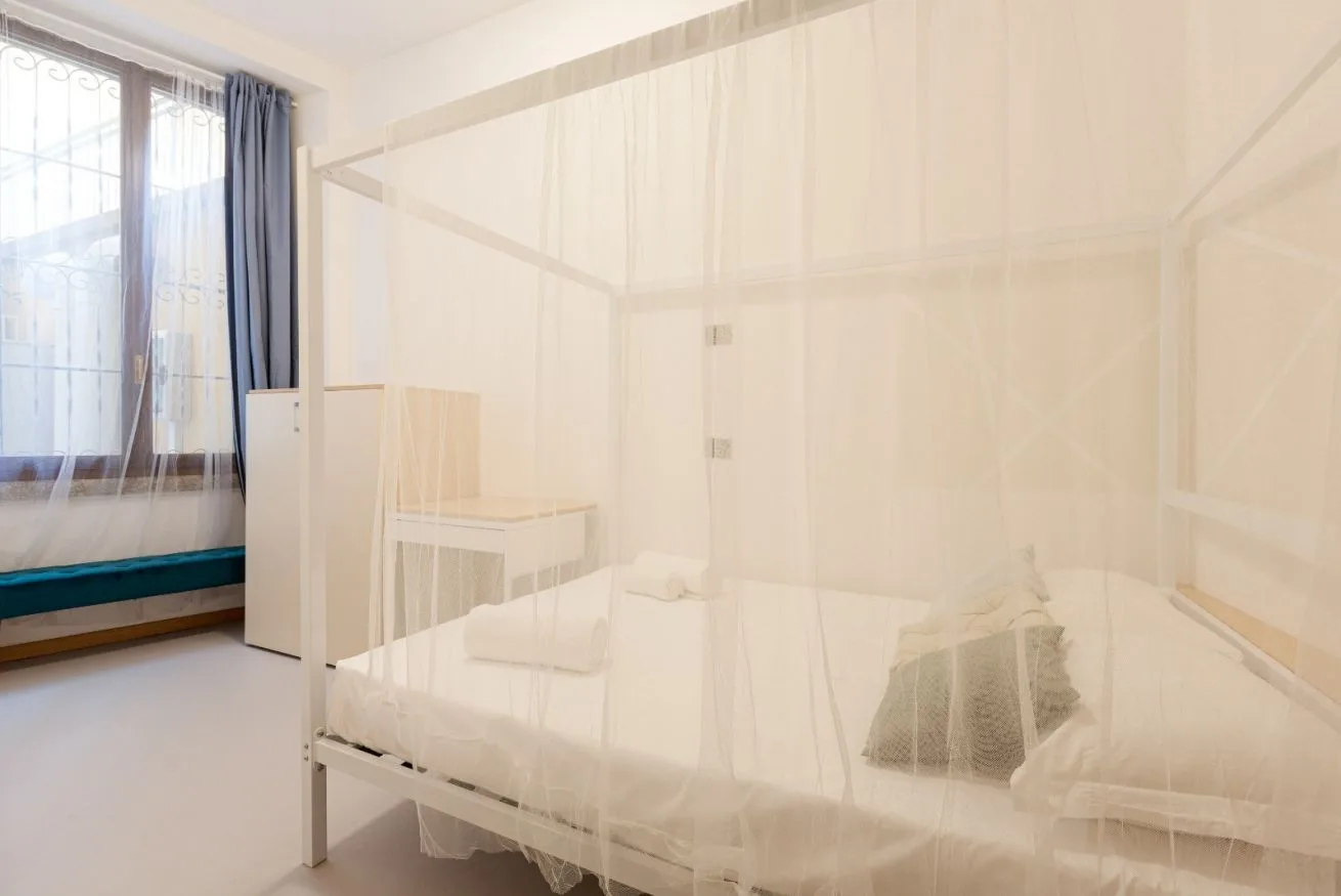 A bedroom with a bed draped in a sheer canopy beside a large window.