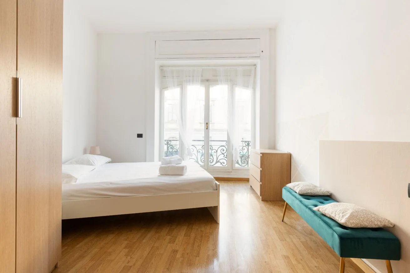 A bright bedroom with a bed, wooden flooring, and a tall glazed door.