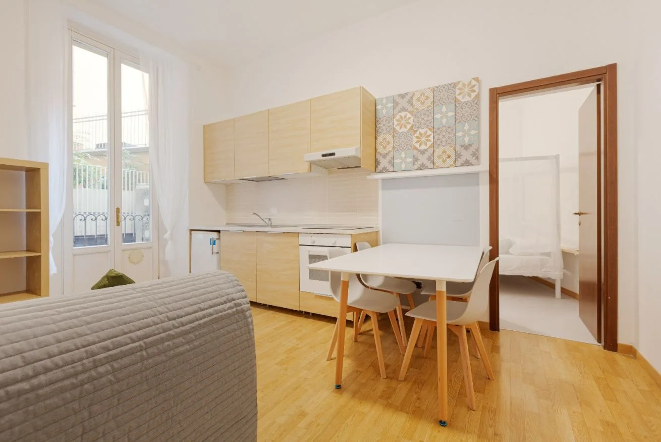 A bright room with a small kitchen, dining table, and doorway to a bedroom.
