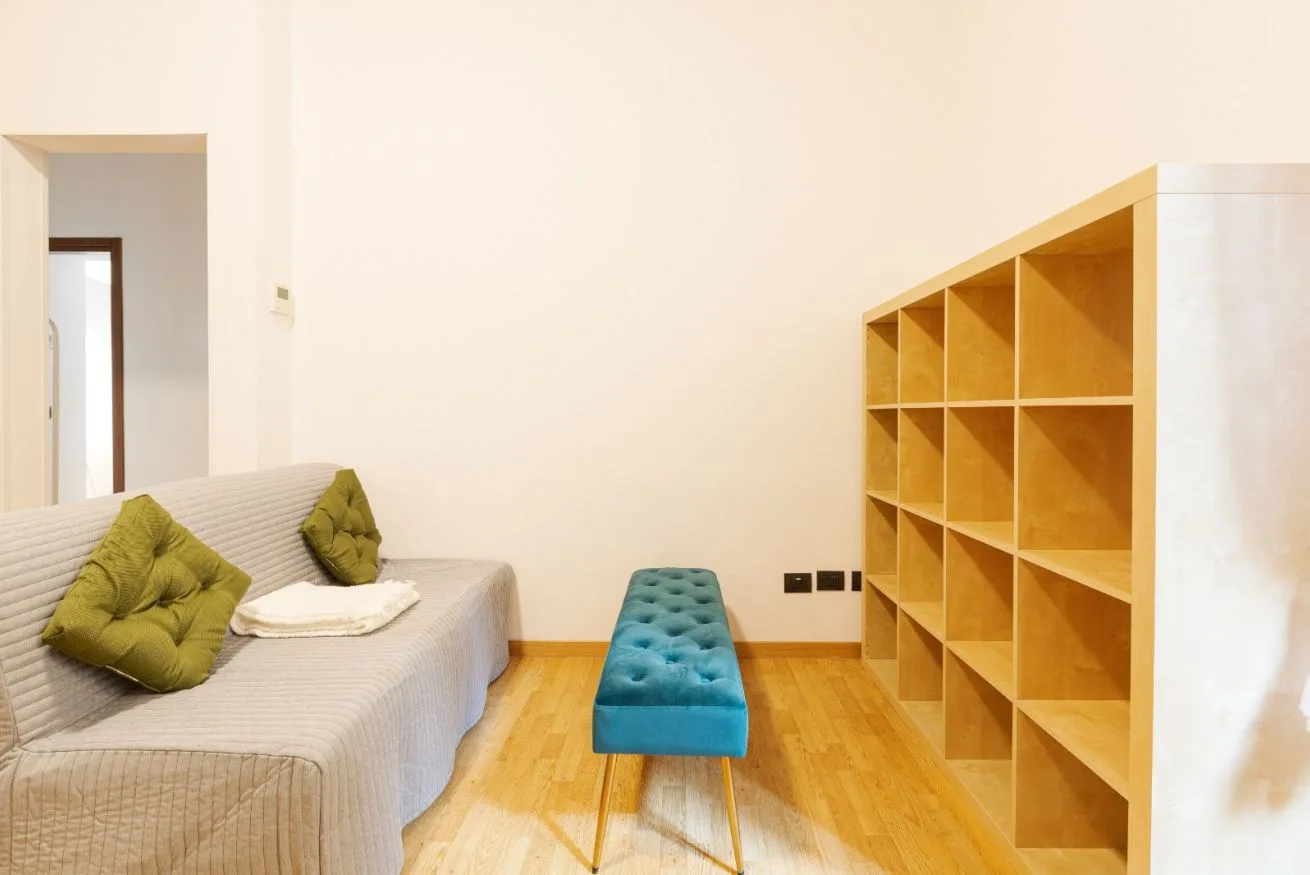 A bright room with a gray bed, turquoise bench, and wooden shelving.