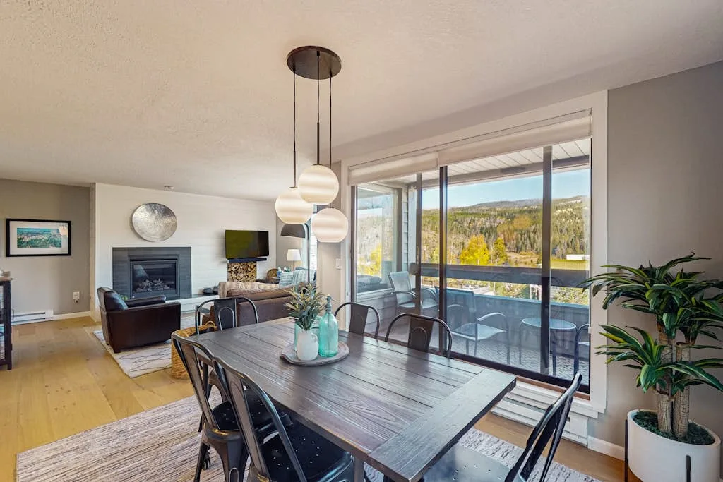 A dining area with a wooden table beside large windows overlooking trees.