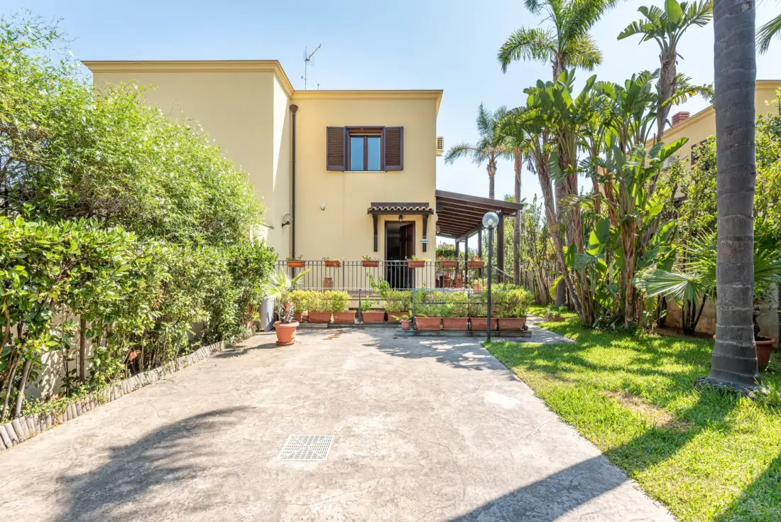Una casa amarilla está junto a una entrada pavimentada y un jardín con palmeras.