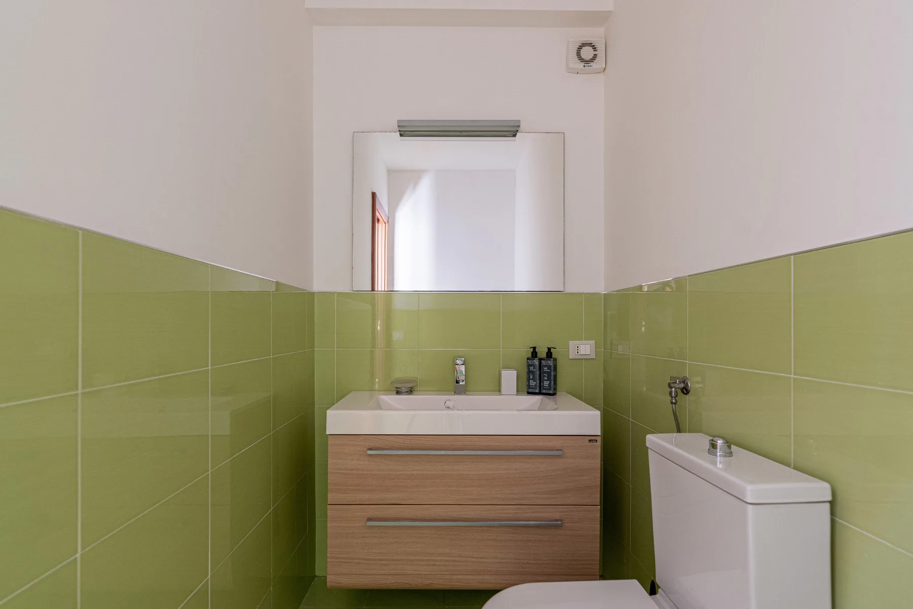 Un baño con mueble de lavabo e inodoro, con azulejos verdes en las paredes.