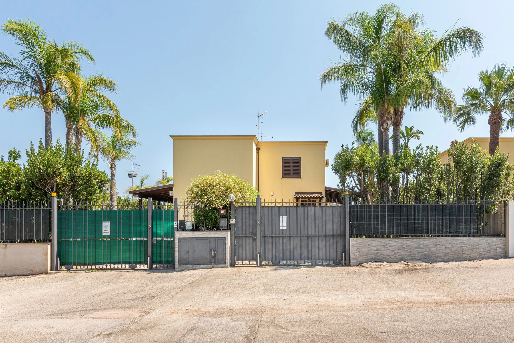 Una casa amarilla está detrás de una entrada con portón, rodeada de palmeras.