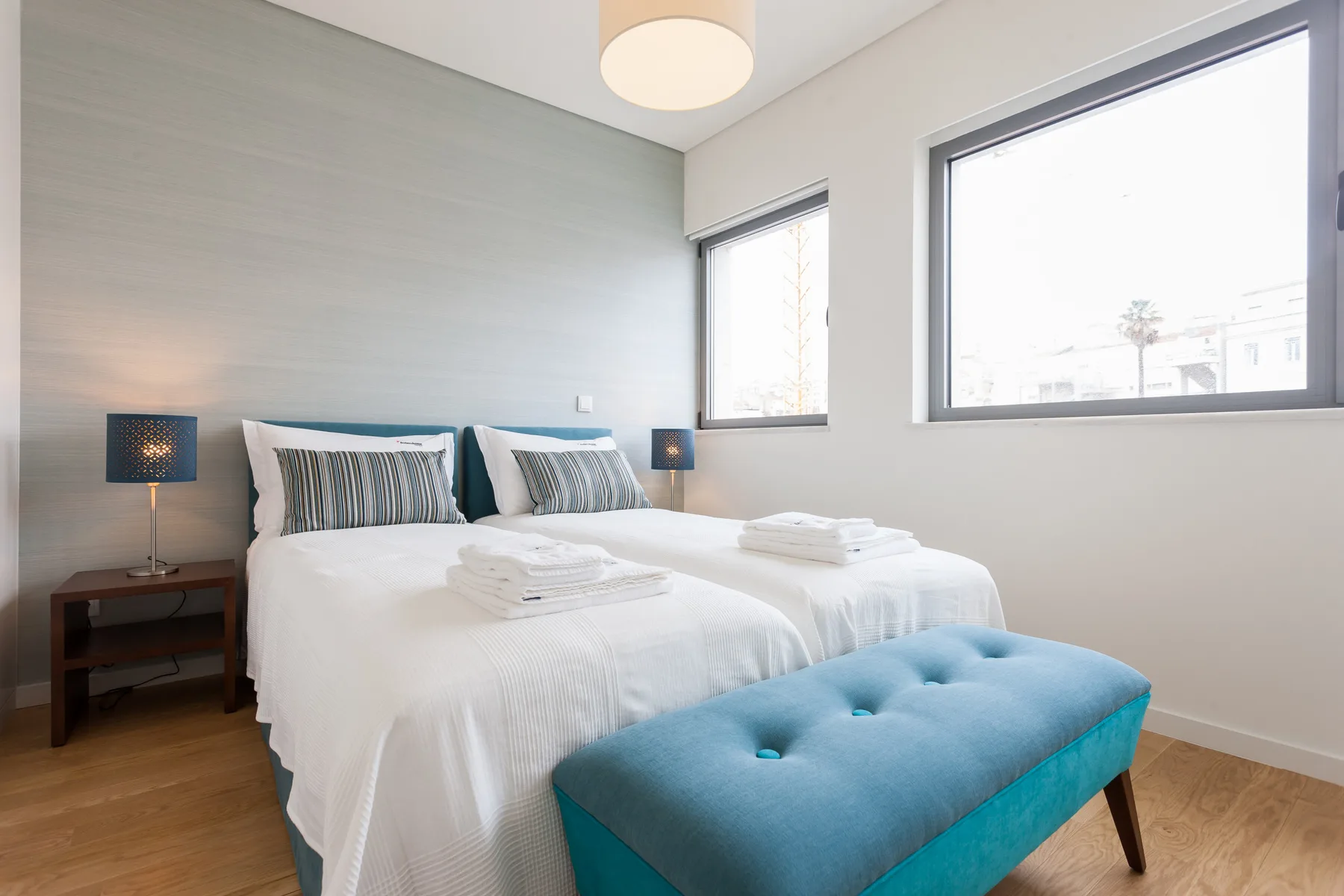 Un dormitorio luminoso con una cama, mesitas de noche y un banco azul tapizado.