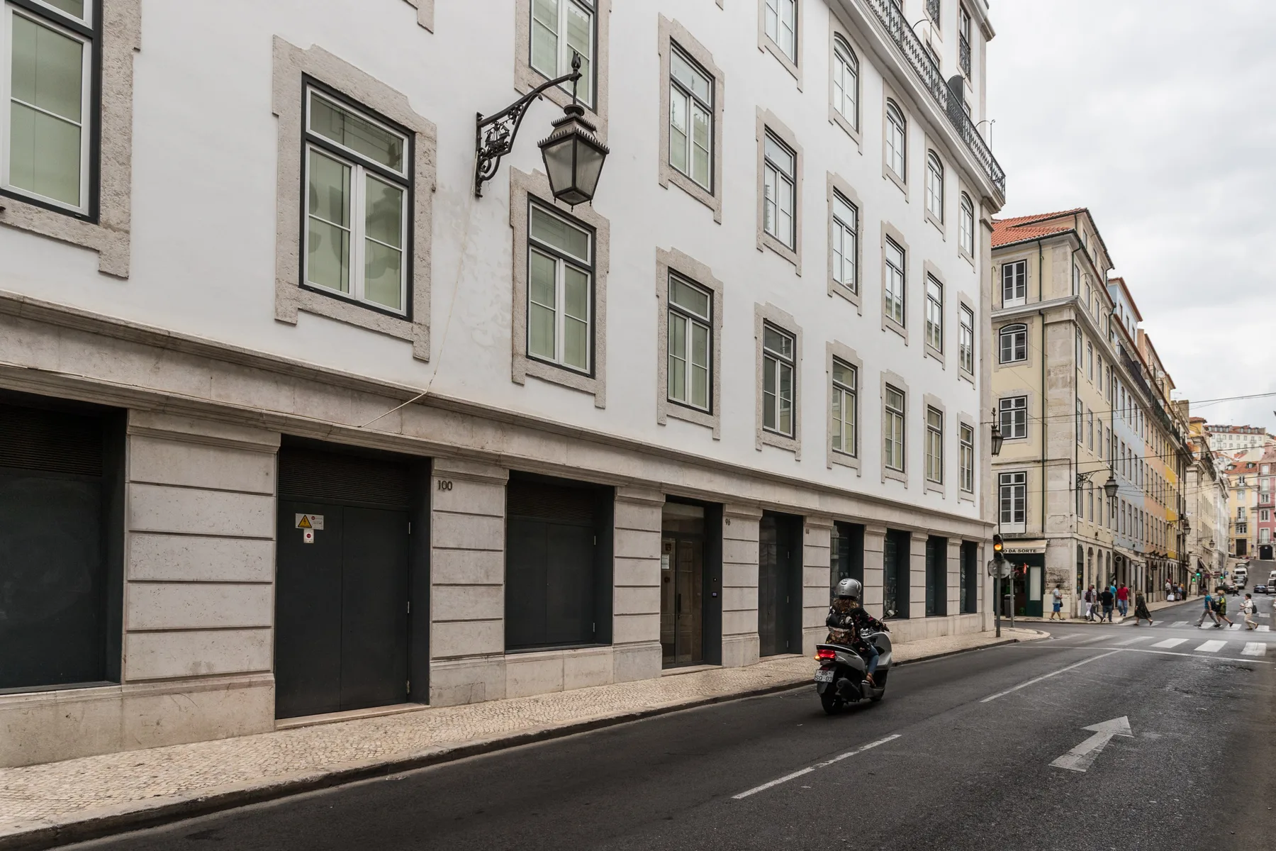 A city street runs alongside a row of pale buildings, with a scooter on the road.