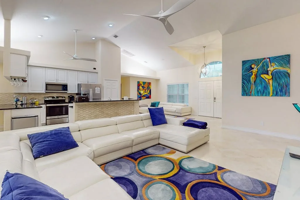 An open-plan living room with white seating, a colorful rug, and an adjoining kitchen.