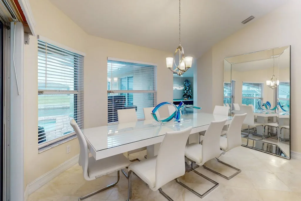 A bright dining area features a long table, white seating, and large windows.