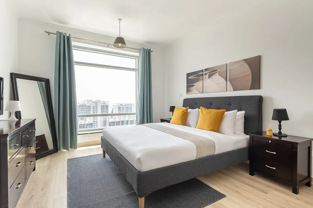 Un dormitorio luminoso con una cama bien arreglada y una gran ventana con cortinas verde azulado.