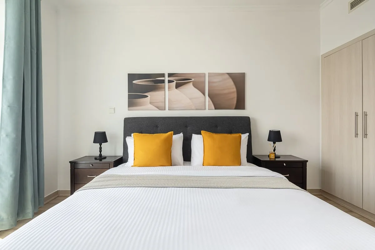 Un dormitorio con una cama bien tendida, cojines amarillos y mesitas de noche.