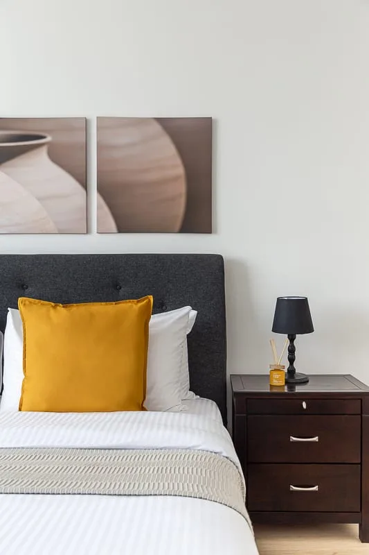 Un dormitorio con una cama bien tendida, un cojín mostaza y una mesita de noche.