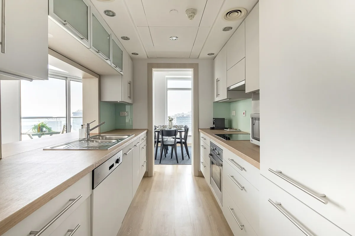 Una cocina alargada y luminosa con gabinetes blancos y una mesa junto a la ventana.