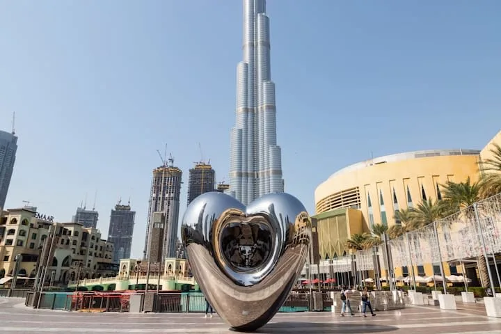 Una escultura metálica con forma de corazón está en una plaza, con el Burj Khalifa al fondo.