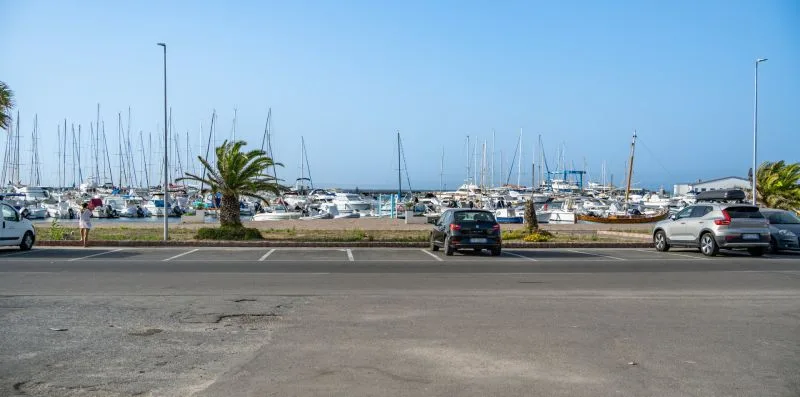 Una marina llena de veleros se ve más allá de una zona de estacionamiento pavimentada.