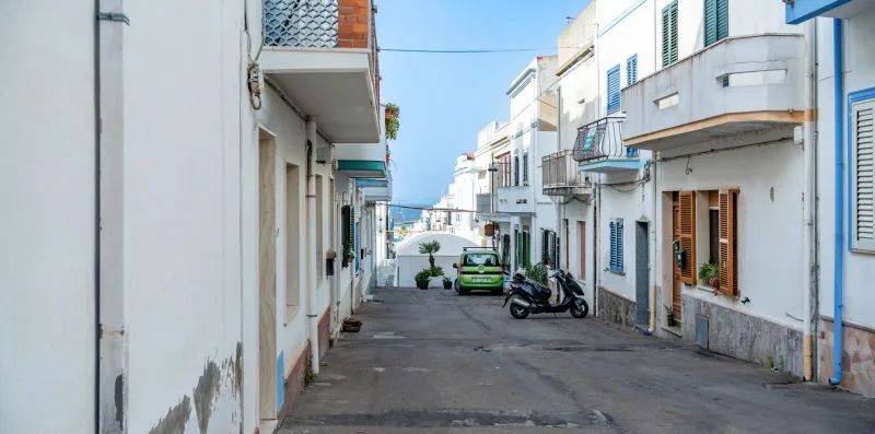 Una calle pavimentada y estrecha pasa entre edificios blancos, con un coche y scooters estacionados.