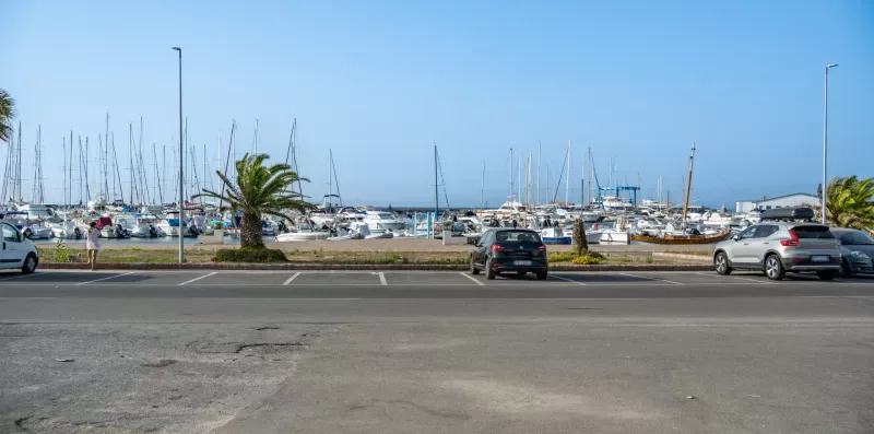 Se ve una marina con veleros más allá de una zona de estacionamiento pavimentada.