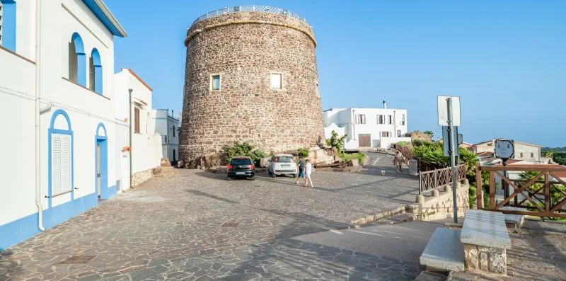 Una torre redonda de piedra se alza junto a una plaza pavimentada con coches estacionados.