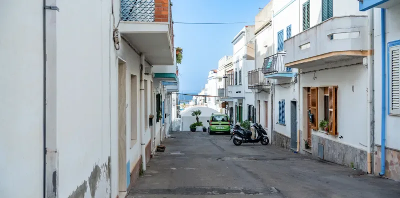 Una calle estrecha bordeada de edificios blancos, con un coche y motos estacionados.