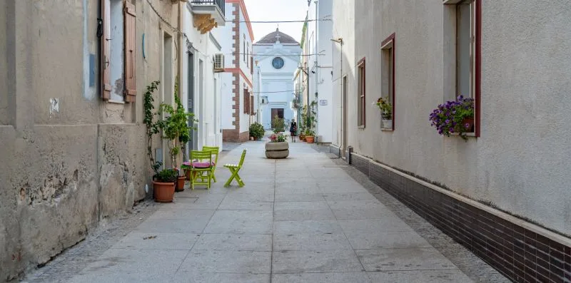 Una calle estrecha y pavimentada entre edificios, con asientos al aire libre y plantas en macetas.