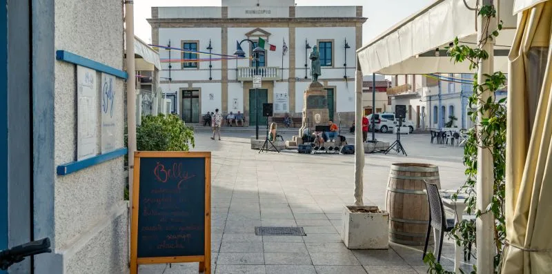 Una plaza con un monumento, mesas de café y edificios alrededor.