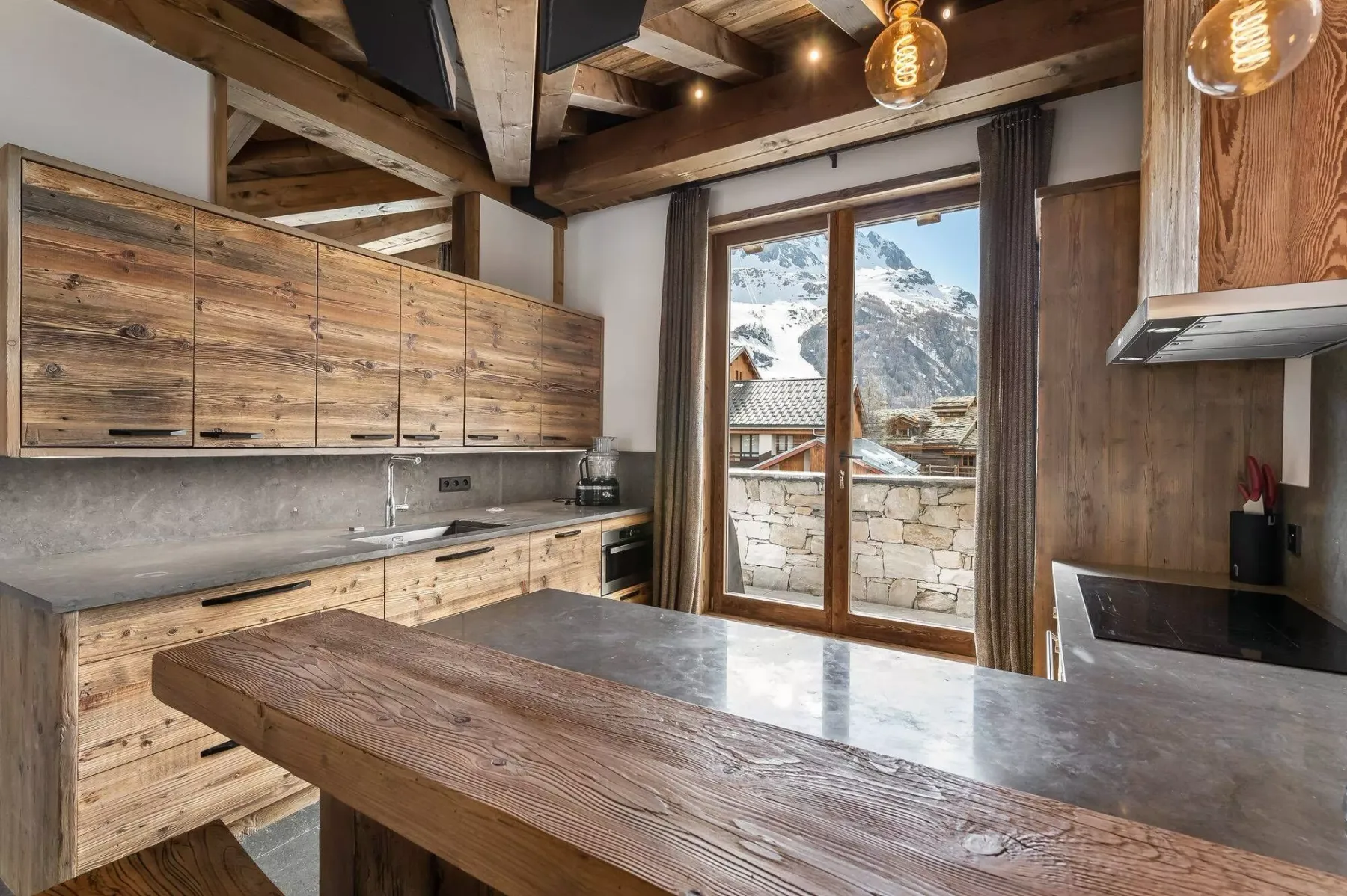 A rustic kitchen with wooden cabinets, a broad island, and mountain views through glass doors.