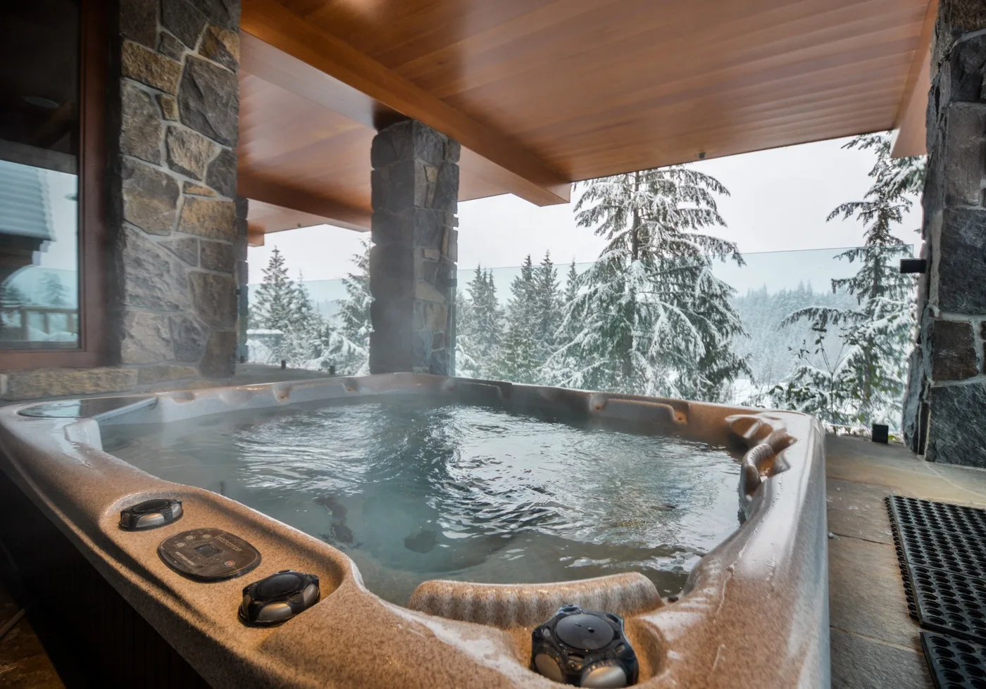 A steaming hot tub sits on a covered deck overlooking snow-covered trees.