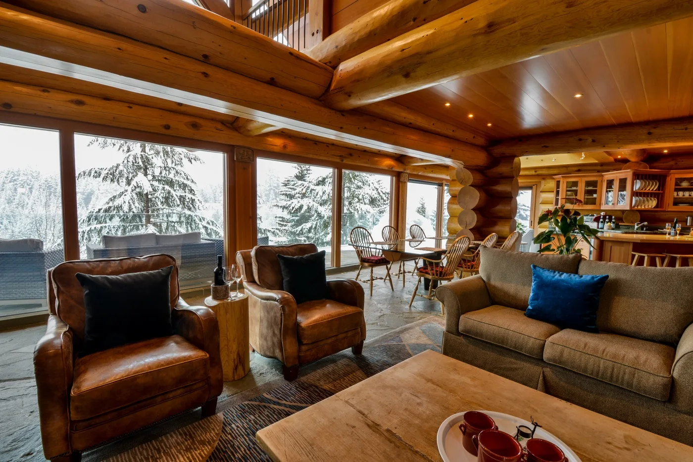 Una sala de estar de cabaña de troncos con asientos junto a grandes ventanas con vistas a árboles nevados.