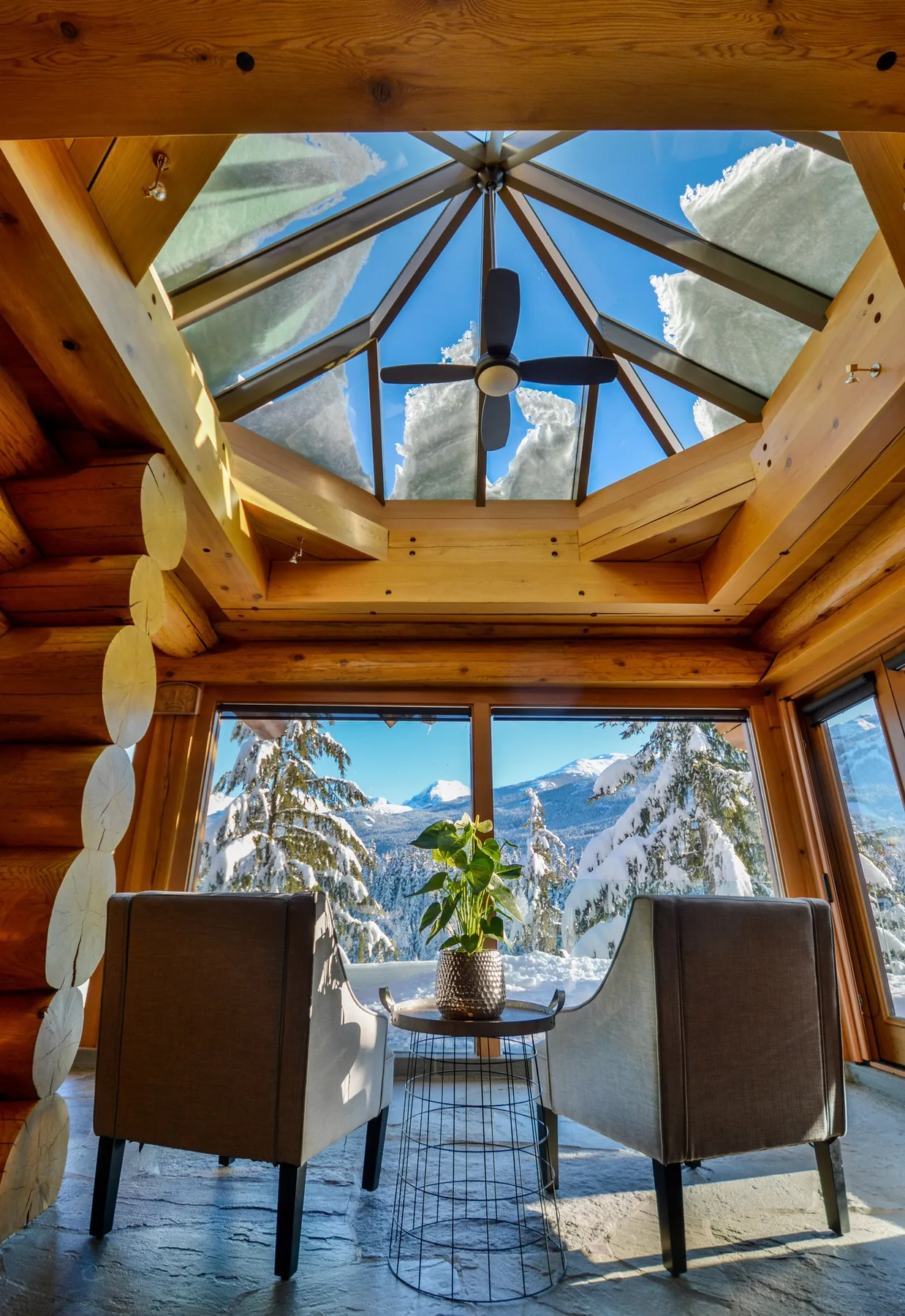 A bright timber room has seating beneath a glass ceiling, with snowy mountains beyond the windows.