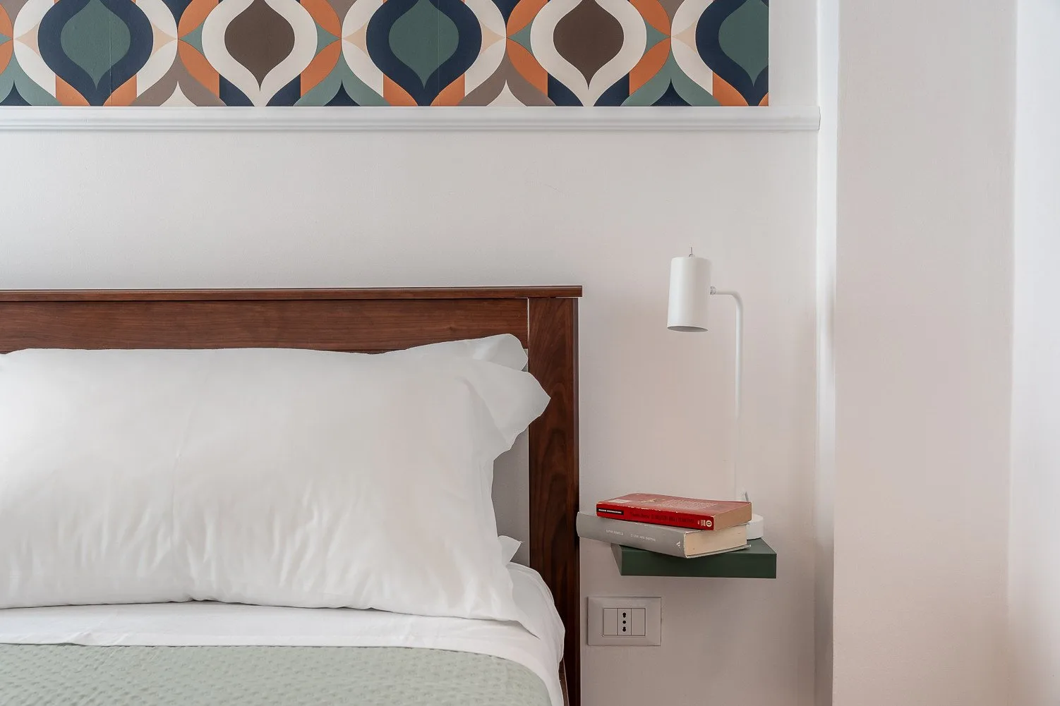 Un dormitorio con cabecero de madera, ropa de cama blanca y una pequeña repisa junto a la cama.