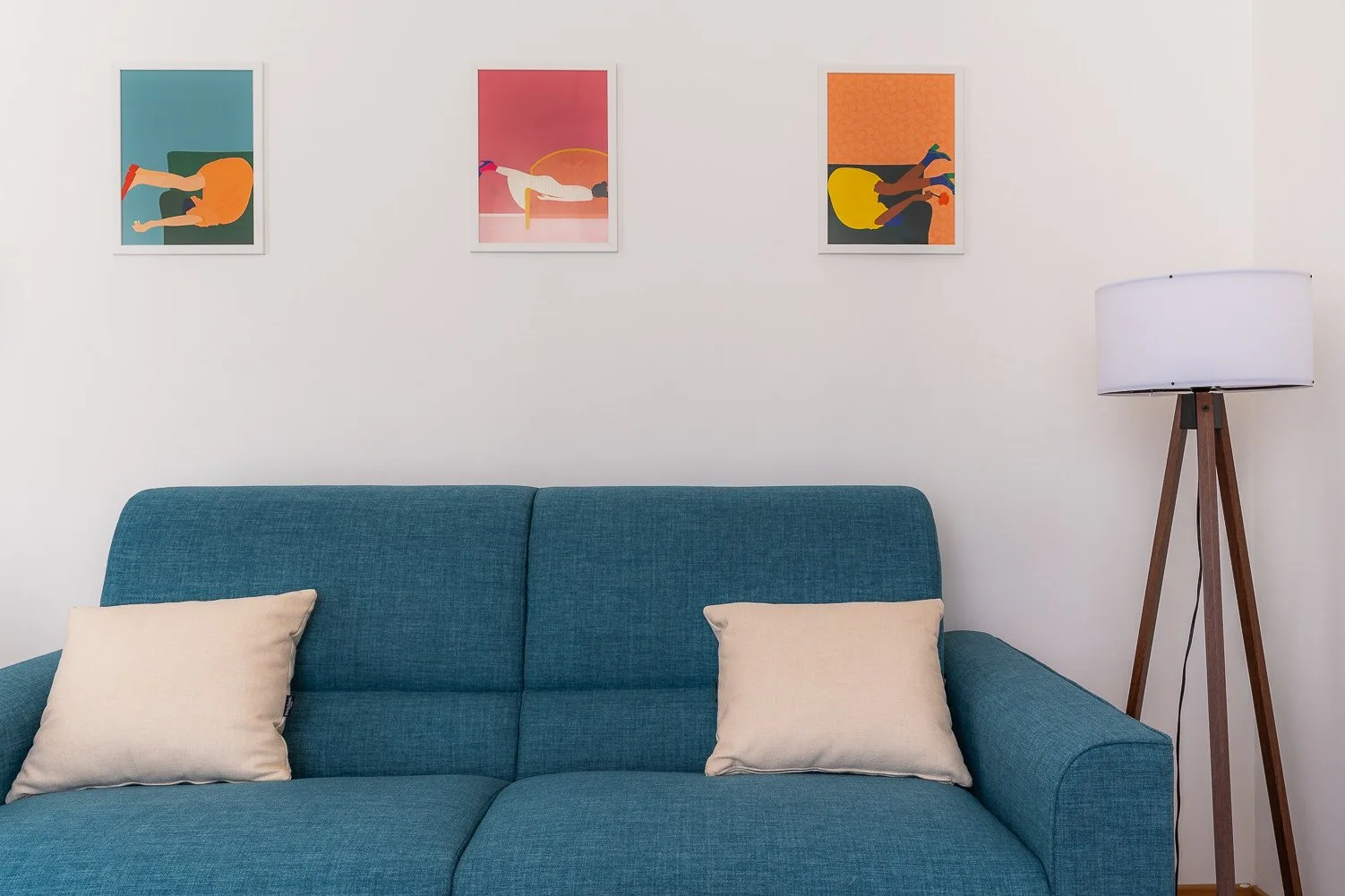 A living room with a teal sofa, light cushions, and colorful framed prints.