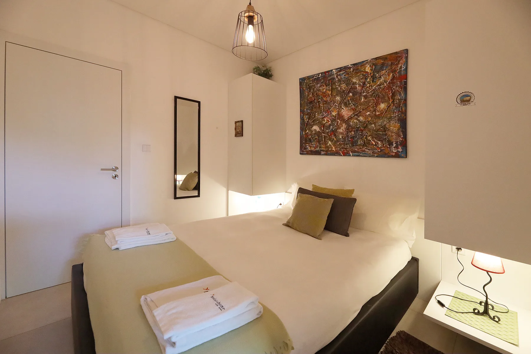 A bedroom with a neatly made bed, folded towels, and colorful wall art.