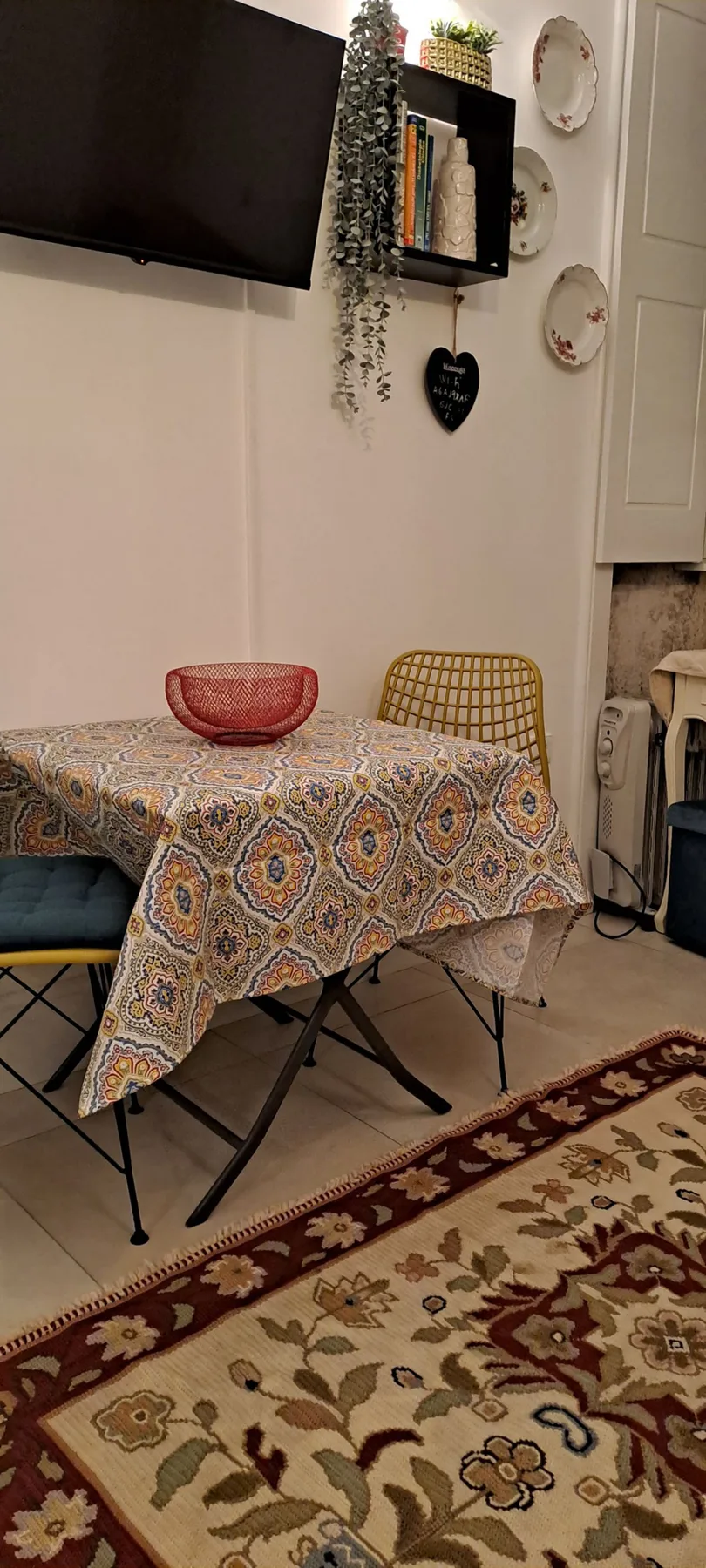 A dining area with a patterned tablecloth, wall-mounted television, and small shelves.