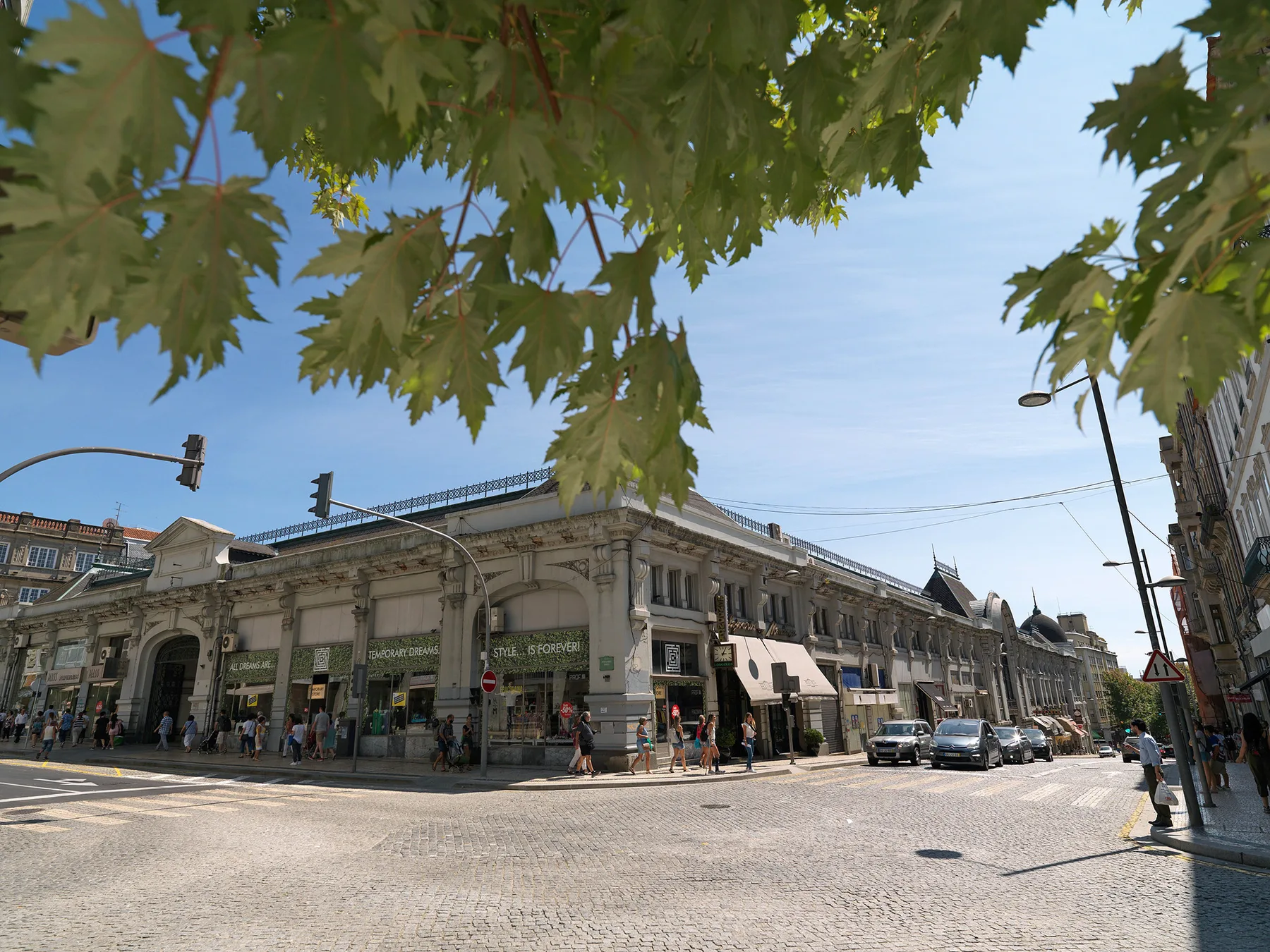 A city street corner with a long stone-faced building and leafy branches overhead.