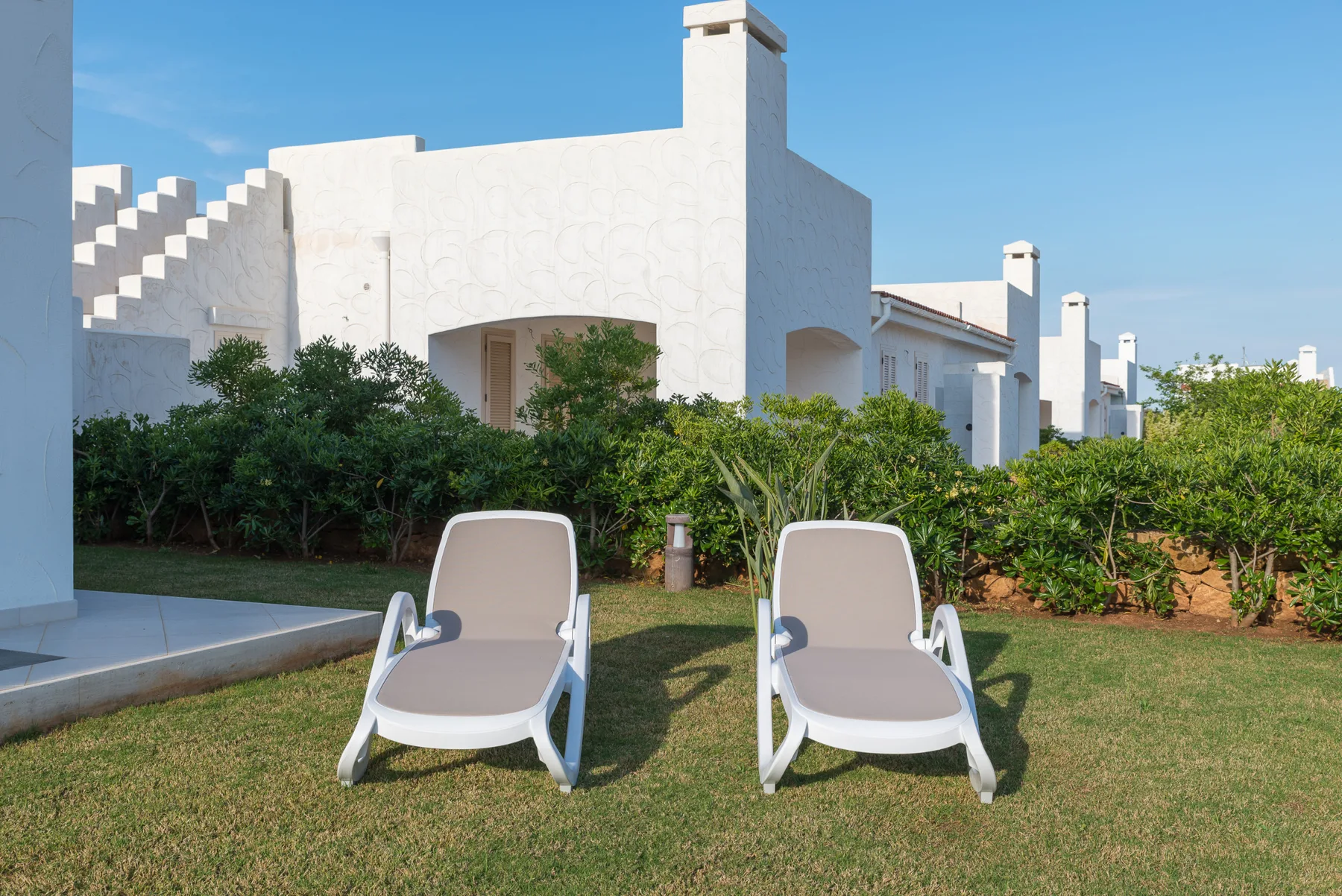 Two outdoor lounge chairs sit on a lawn in front of a white building and greenery.