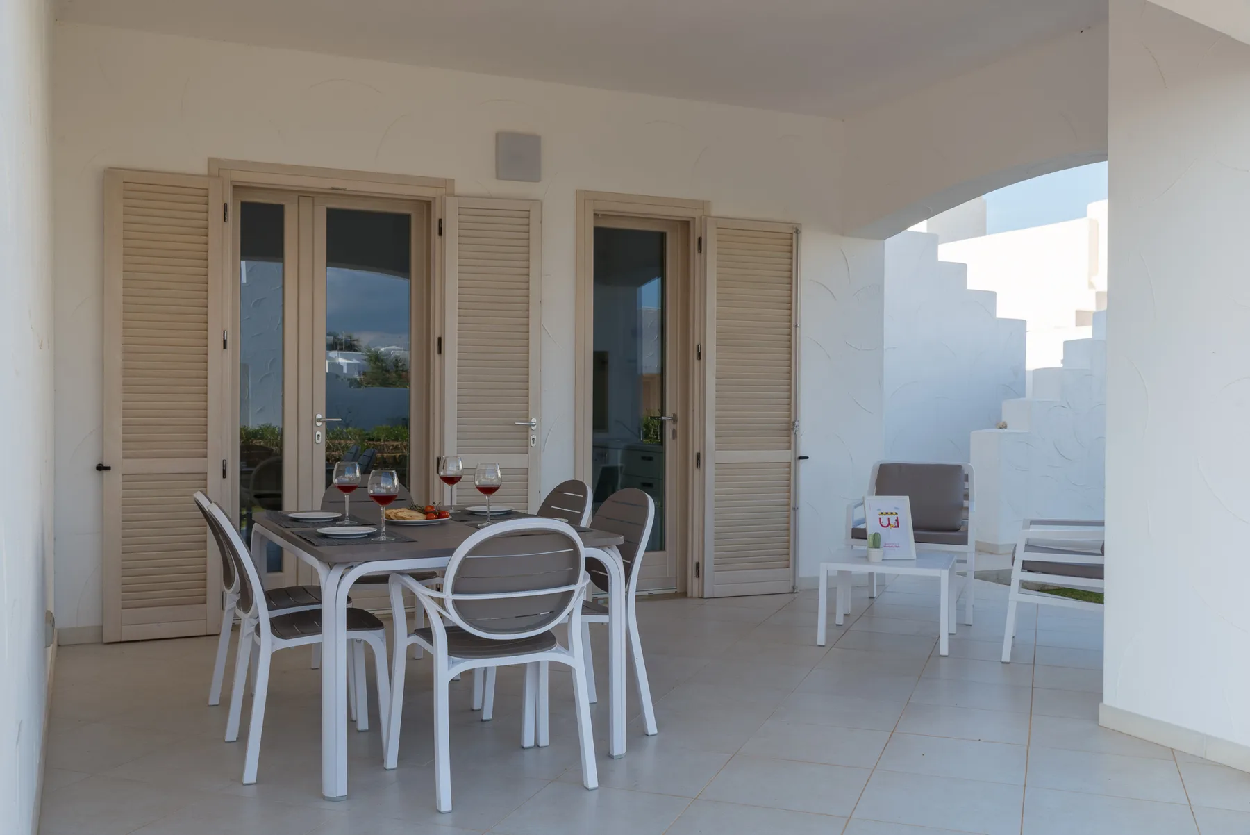 A covered patio with a dining table and white seating near open shuttered doors.
