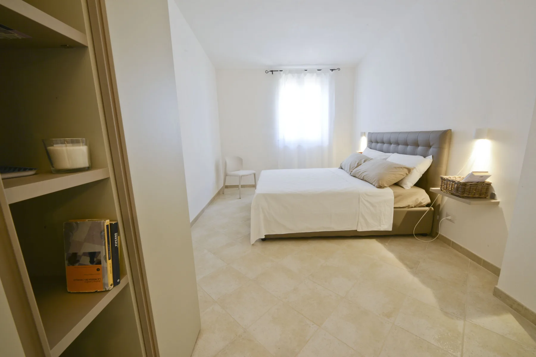 A bedroom with a bed, a chair, and shelving beside a bright window.