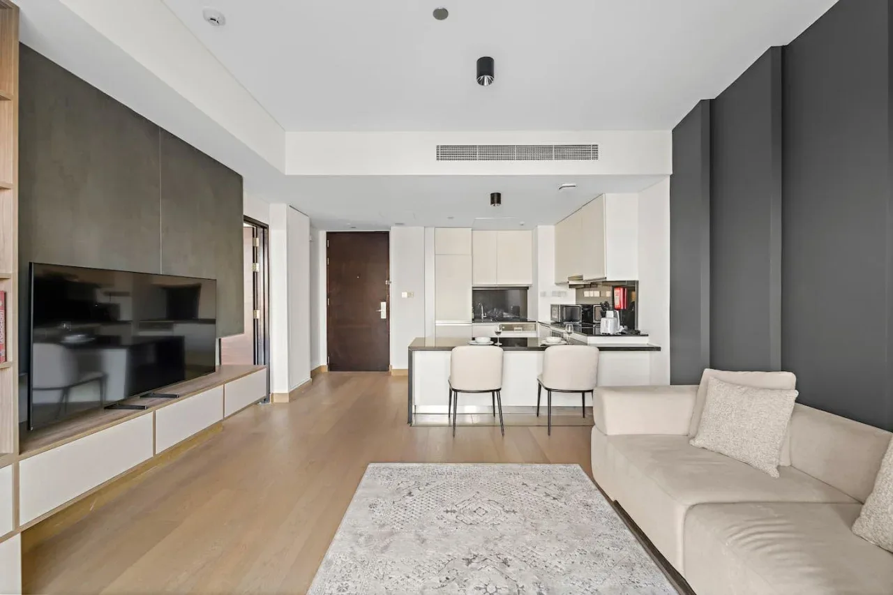 A modern living room opens onto a kitchen, with a television and light-colored seating.