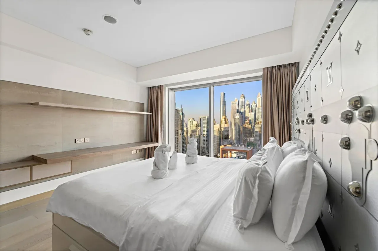 A bedroom with a neatly made bed and large windows overlooking city buildings.