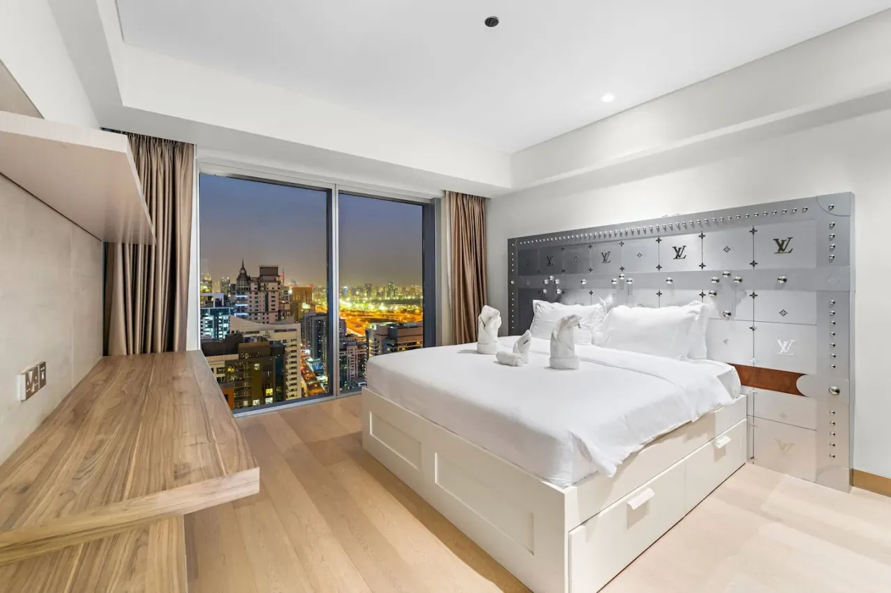 A bedroom with a large bed beside floor-to-ceiling windows overlooking city lights.